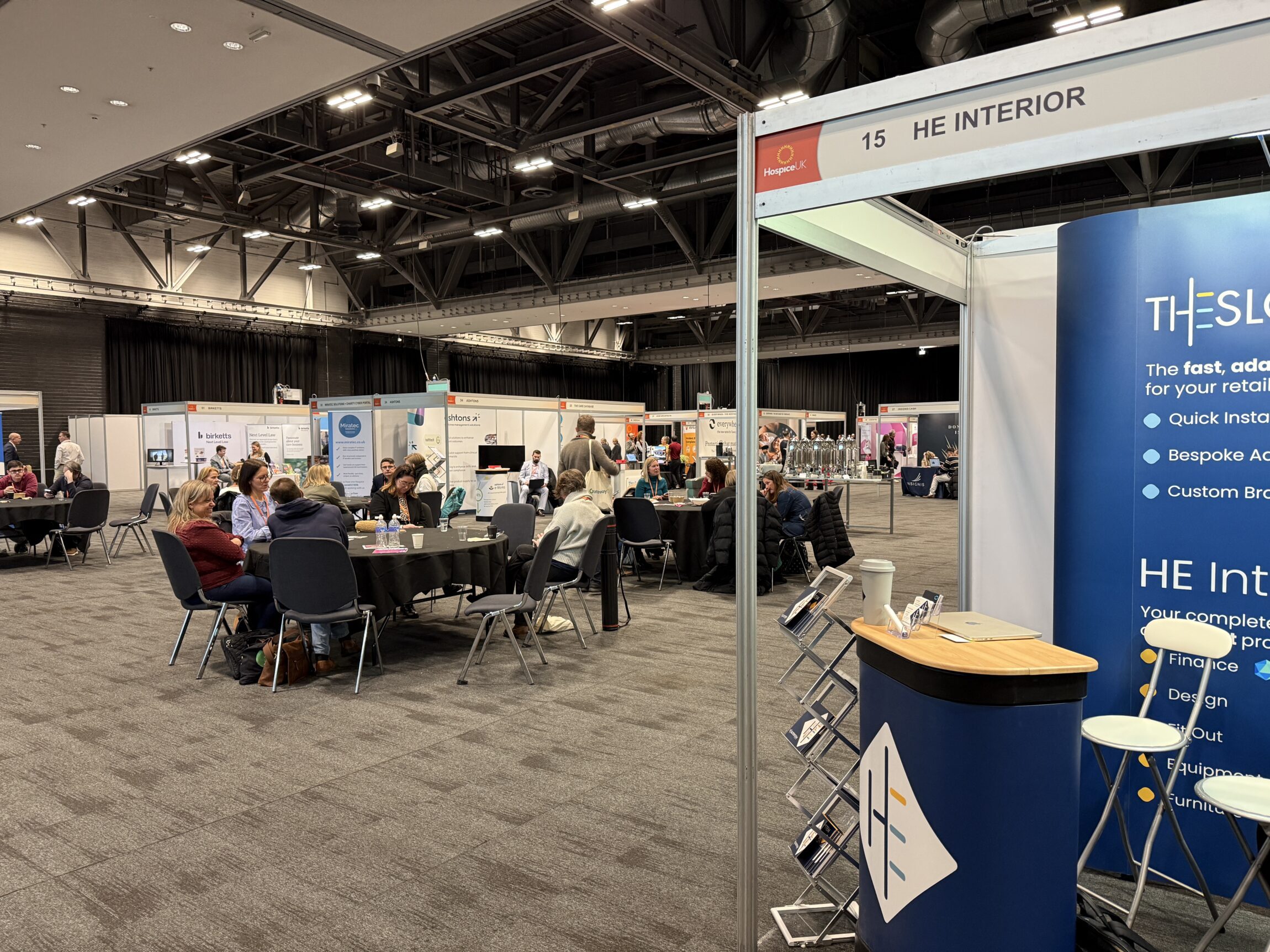 HE Interiors exhibition stand at the Hospice UK National Conference, with attendees seated at tables and visiting stalls in a large conference hall.