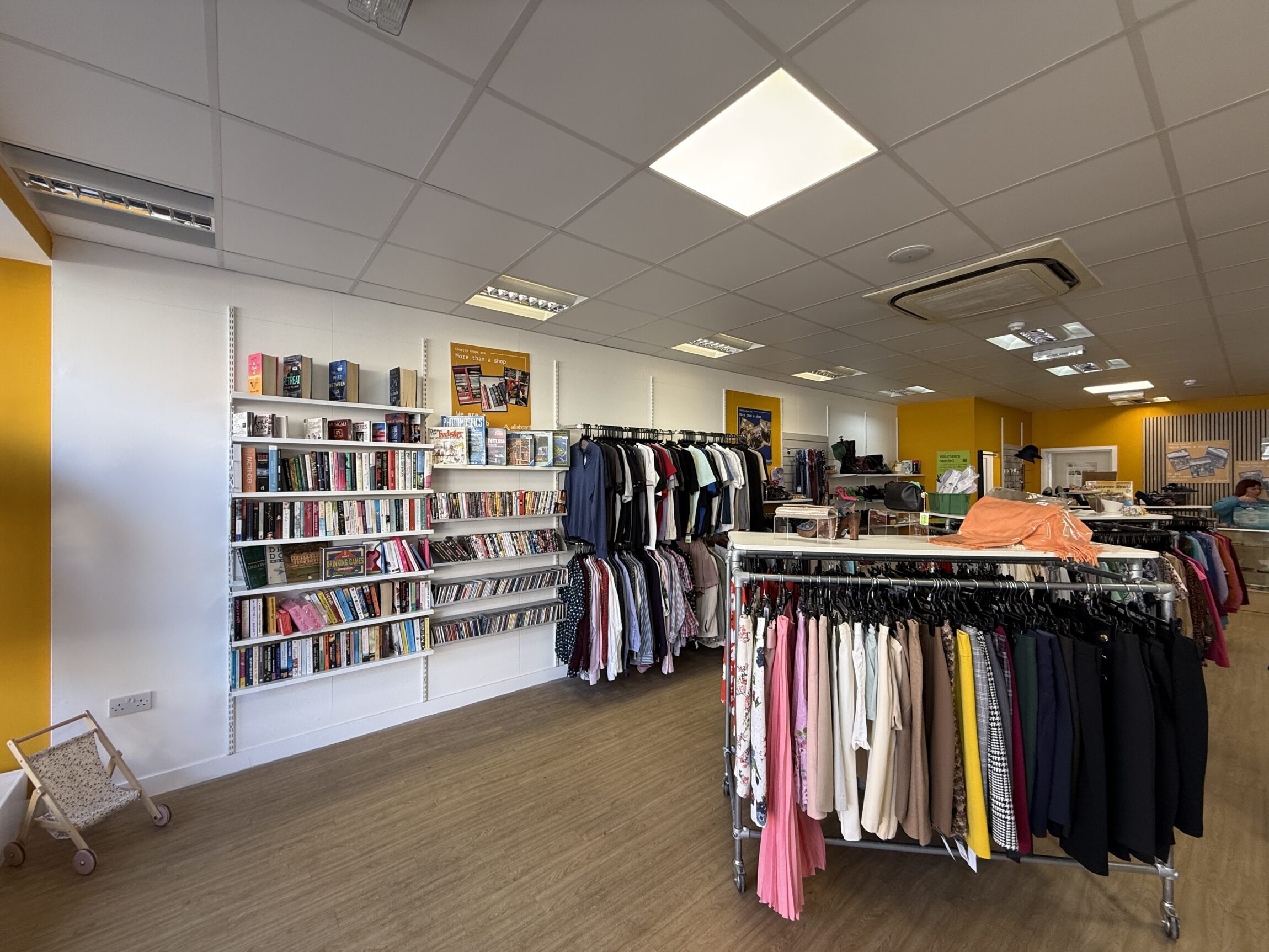 Interior of All Aboard charity shop in Fleet showing clothing rails, shelving with books, DVDs, and board games, and bright yellow feature walls.