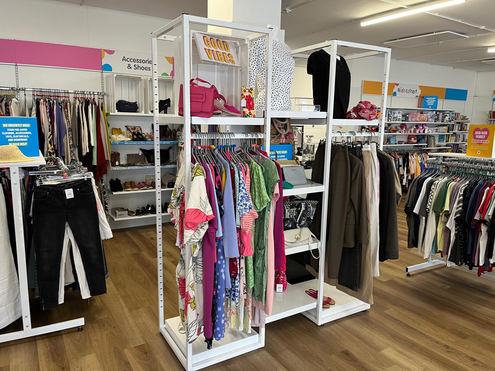 Bright, modern charity shop interior with white modular clothing display units, neatly arranged garments, accessories, and wooden flooring.