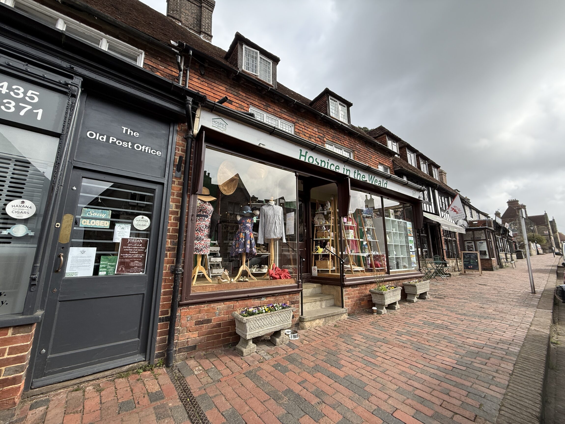 Exterior of Hospice in the Weald charity shop on a historic high street in Mayfield, showing a brick façade, large display windows with mannequins, and a cobbled pavement.