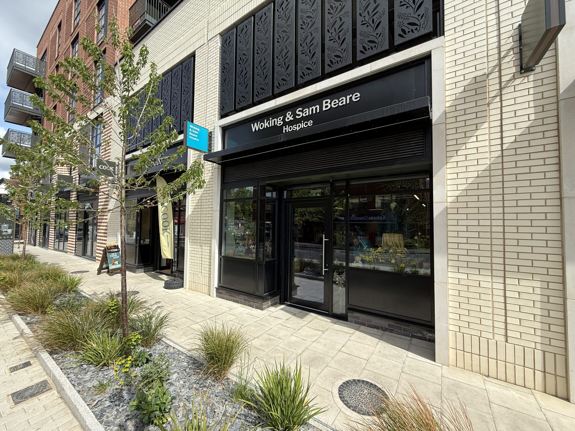 Front exterior of Woking & Sam Beare Hospice charity shop in West Byfleet, showing a modern storefront with black signage, large windows, and landscaped pavement in a new development.