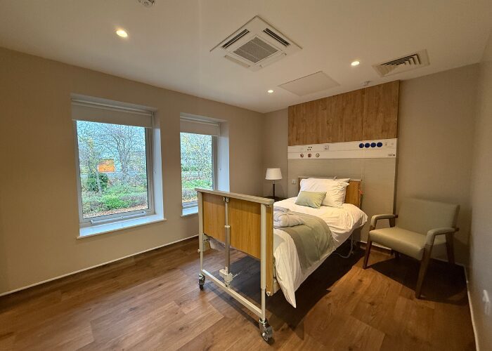 Modern private clinic patient room with a hospital bed, wooden feature wall, clinical fittings, armchairs and natural light from two windows designed by HE Interiors with their turnkey clinic service.